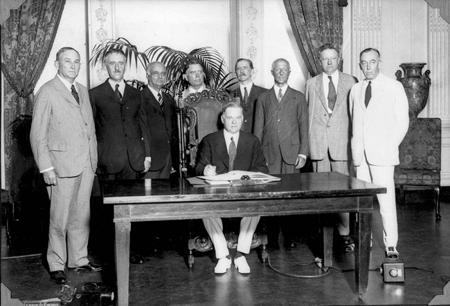 A group of men stand around a seated Herbert Hoover who poses as he signs a treaty.