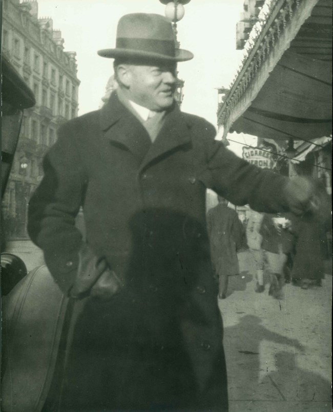 A man in a hat and overcoat walks a city street.