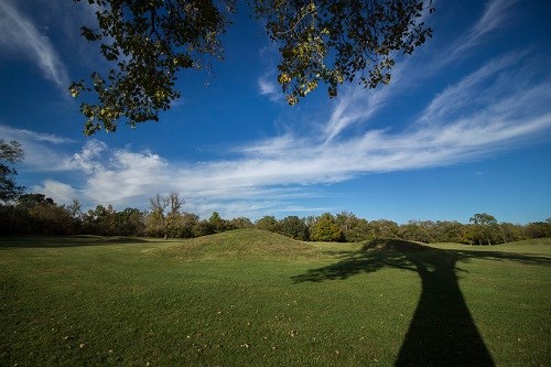 Grass-covered mounds surrounded by trees under a blue sky