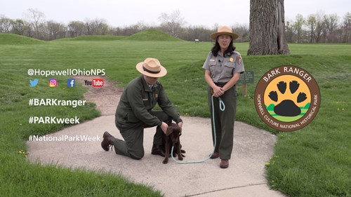 Two park rangers wearing tan flat hats with a brown dog stand on a concrete pad surrounded by green grass and earthen mounds. BARK ranger logo on right. Text on left reads @HopewellOhioNPS, #BARKranger, #PARKweek, #NationalParkWeek