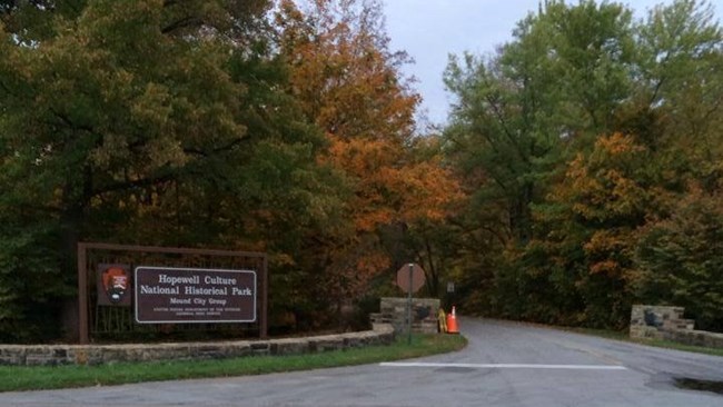 Several multi-colored trees behind a low stone wall and a large wooden park entrance sign with a road leading into the park.