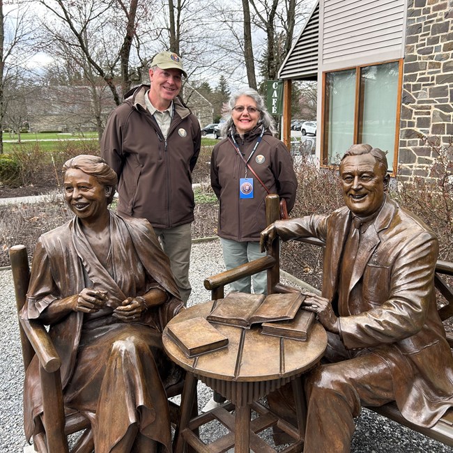 Two smiling NPSvolunteers stand behind bronze statues of Eleanor and Franklin Roosevelt. The volunteers wear official, brown jackets for NPS Volunteers.