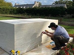 A park ranger cleans a marble block (grave marker) within a garden setting.