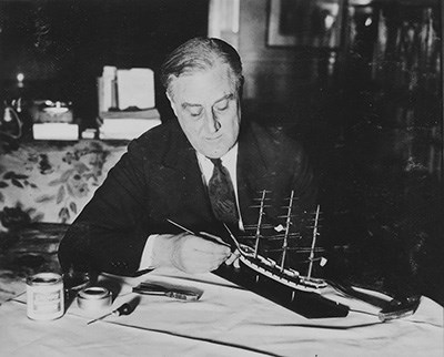 A man (FDR) wearing a suit and tie seated at a desk and working on a ship model.