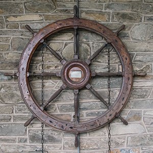A large wooden ship's wheel hanging against a stone wall.