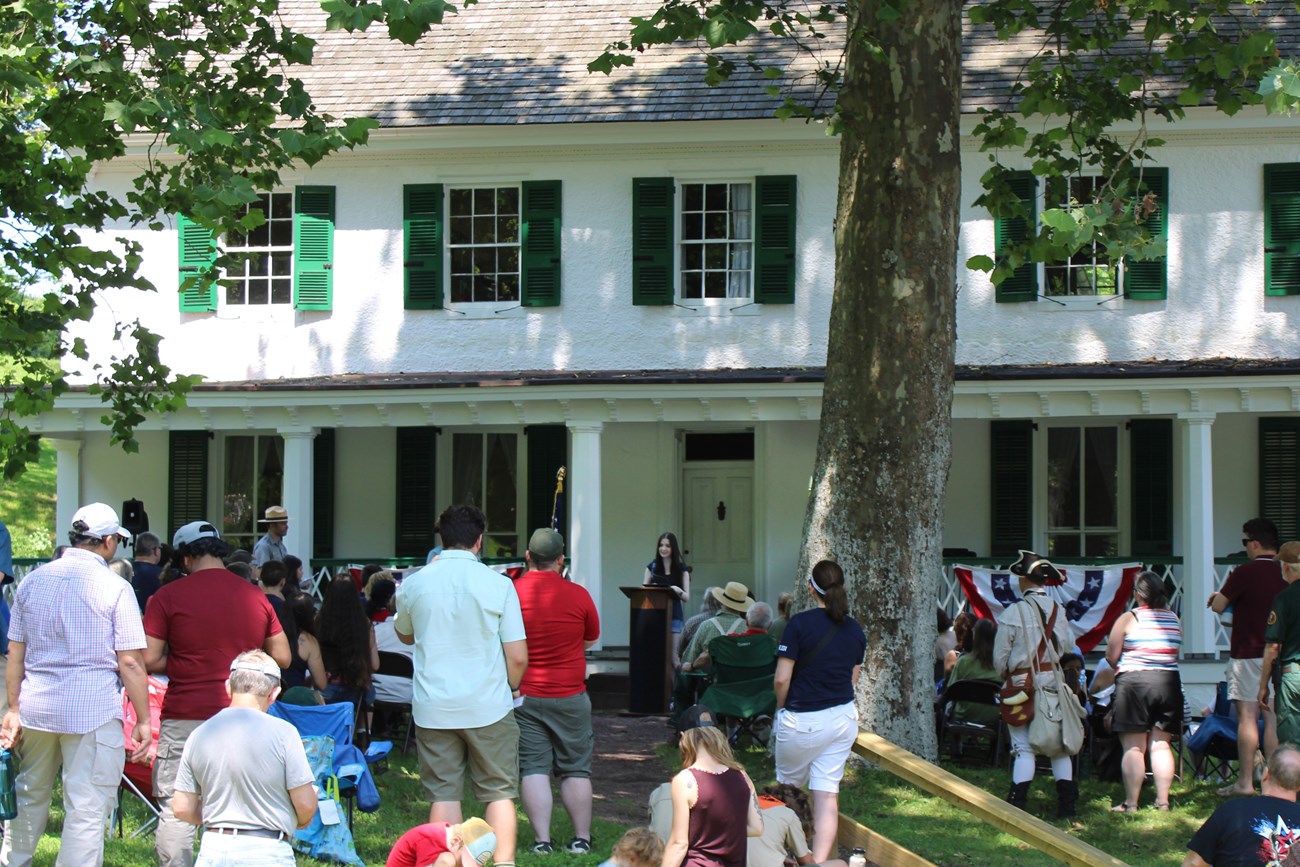 A group of people gather near the front of a large white building. A teenager speaks from the podium to the crowd..