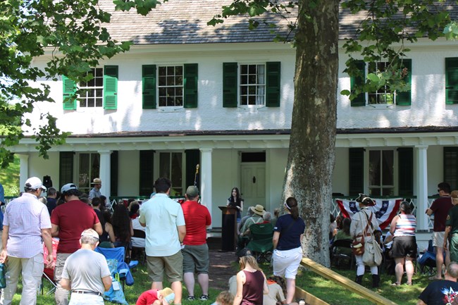 A group of people gather near the front of a large white building. A teenager speaks from the podium to the crowd..