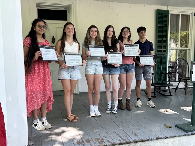 Six teenagers stand next to each other on a porch holding certificates.