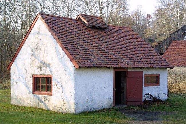 White building with red tile roof. Red door and windows. Door is open. Bits of scrap metal is on side of building.