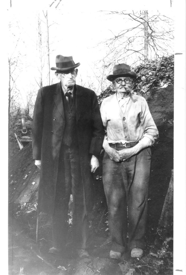 Two men pose for picture by collier hut and charcoal pit.