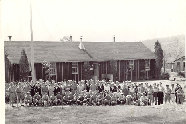 Sailors pose for group photograph. Buildings in background. Sailors all have white service hats.