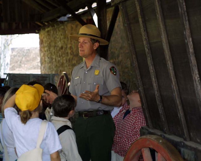 A group of children stand around a park ranger near a wooden large wagon.