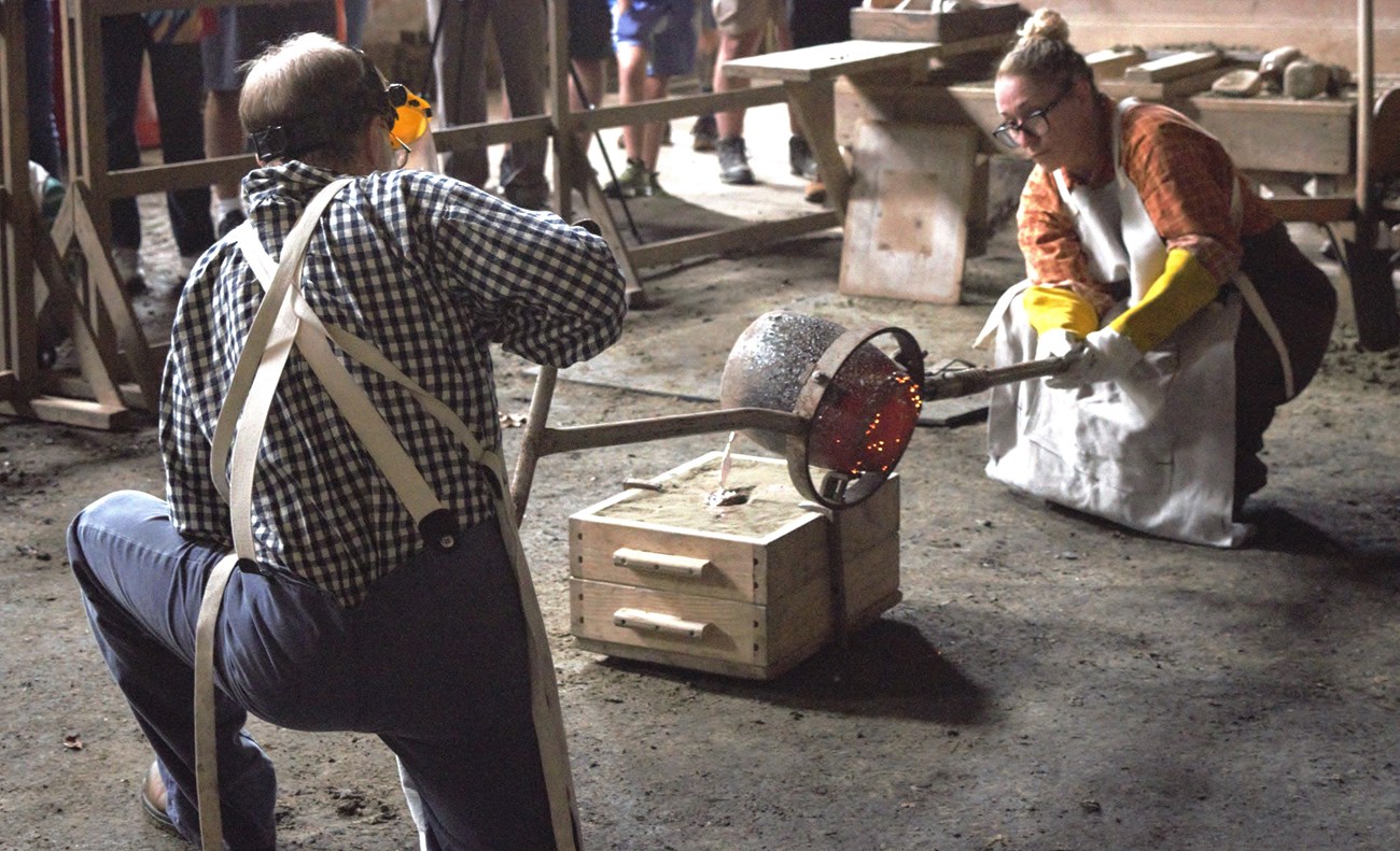 Two people dressed in historical clothing poor mantellic matalic liquid into a mold.