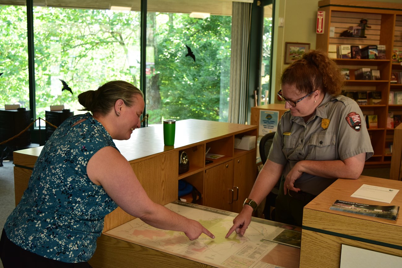 A park ranger talking with a visitor at a desk. They are both pointing at a map.