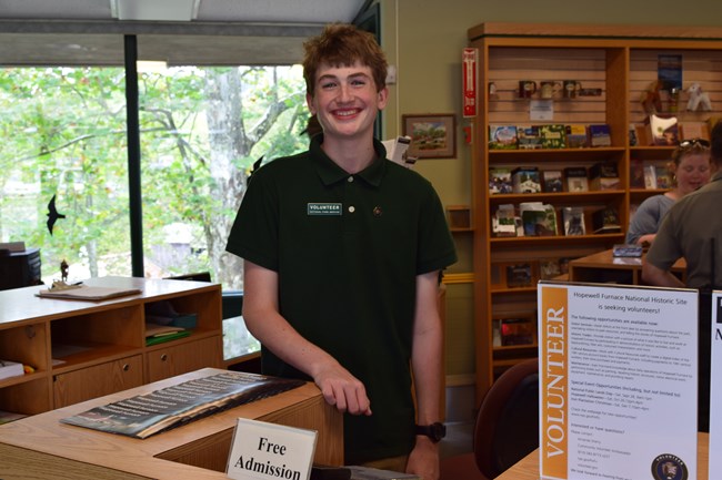 A person smiles at a desk in a store. Their nametag says volunteer. A sign on the desk reads free admission.