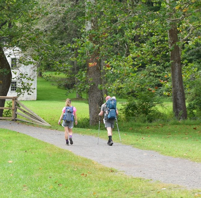 Two people wearing backpacks hike a gravel path. One uses hiking poles.