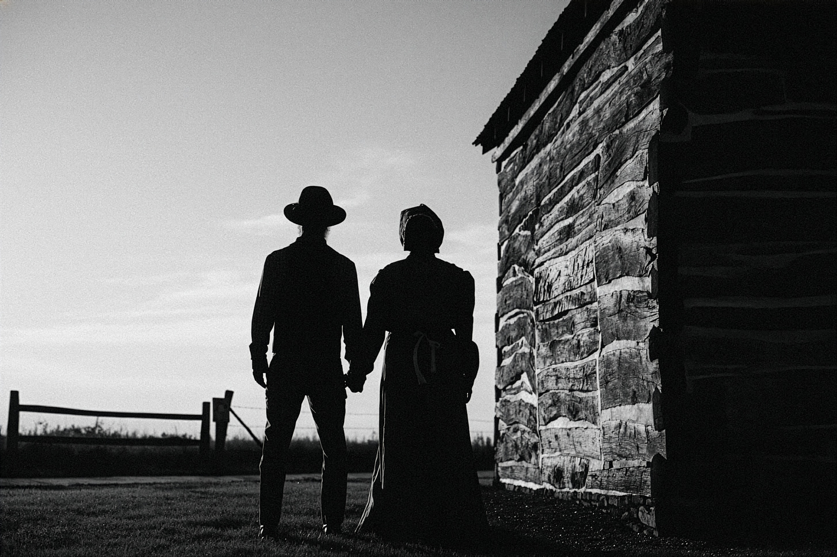 Black and white photo. Two individuals wearing living history clothes are backlit silhouettes next to a log cabin.