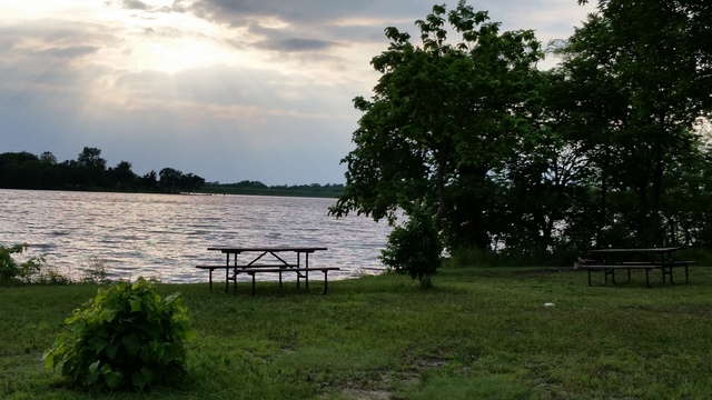 Rockford Lake camping, Sun, low in the sky sends rays over the water. There are trees, green grass, and picnic tables in the foreground.