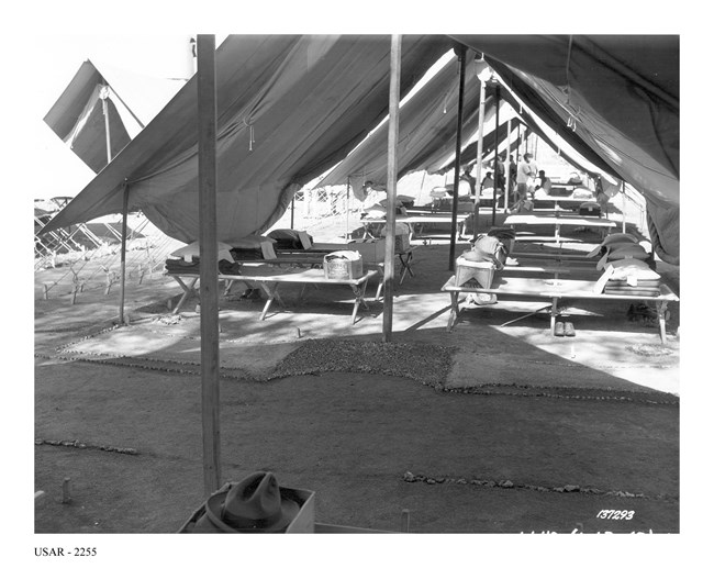 Cots laid out in rows under tents, boxes full of clothes on top of the beds with shoes underneath.