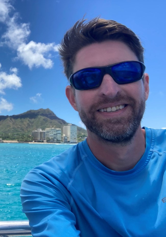a man with short brown hair smiles and wears sunglasses on bright blue waters with an island in the background.