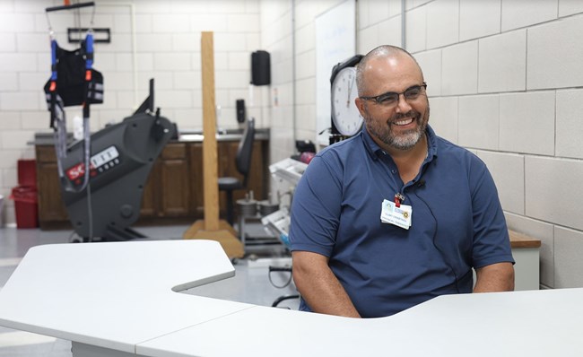 Bald man in blue shirt sits at white desk with exercise equipment behind him.
