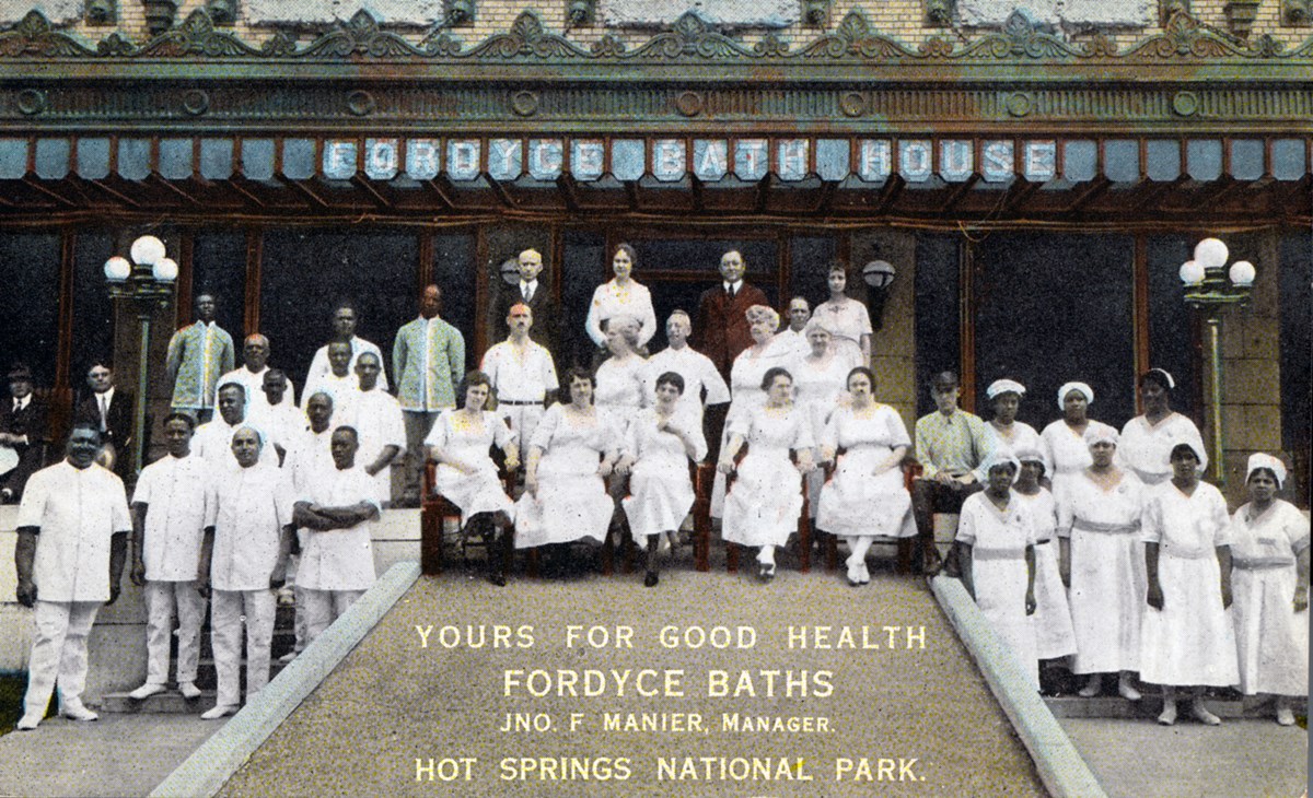 37 men and women stand and sit in front of a building with yellow brick. Most of the men and are wearing all-white uniforms that cover their entire body.