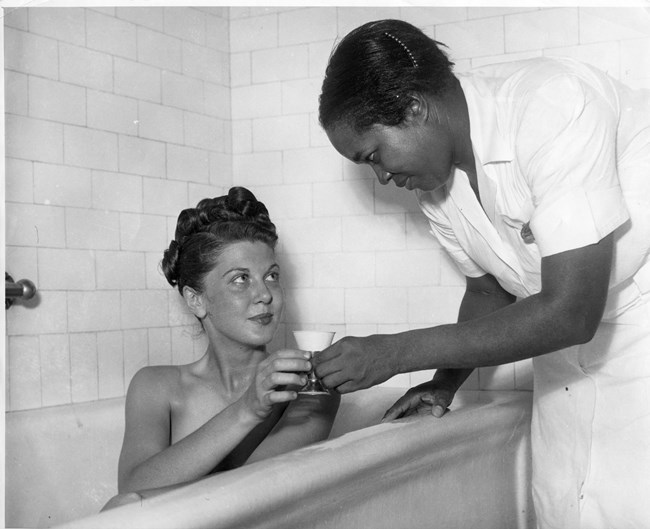 African American woman in white uniform hands a cup to a white woman with dark hair sitting in a bathtub