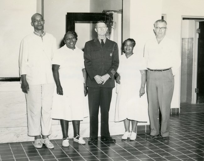 Three men, one African American and two white stand in a line with two African American women. The African American man and women wear white uniforms. One white man wears a white shirt and dark pants. The other wears a dark uniform with a tie.