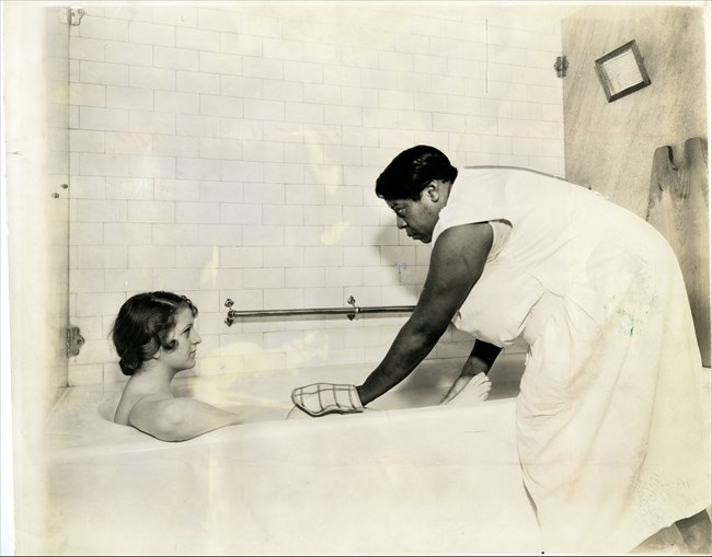 African American woman wears a white uniform and wears a white mitten on her hand. She stands over a bath tub. In the tub is a white woman with dark hair looking up at the African American woman.