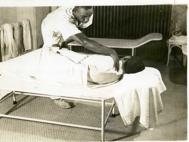 African American man in white uniform rubs the shoulder of white man laying on a metal bed. The white man is wrapped in white towels.