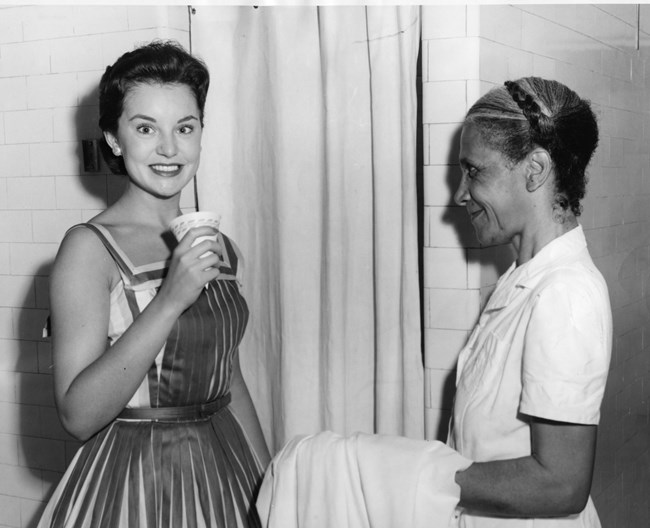 African American woman in white uniform holding white towel stands with white woman with dark hair in white and gray dress holding a cup.