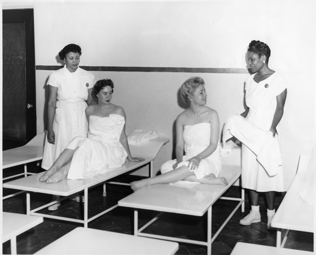 Photo of two African American women and two white women. The African American women are standing wearing white uniforms. One is holding a towel. The white women are sitting on metal beds wearing towels.