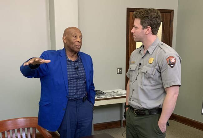 An African American man in a blue shirt, blue jacket, and blue pants speaks to a white man in a gray shirt with gold badge and green pants. They are indoors.