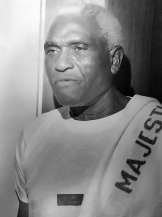 African American man with white hair wears a white shirt. He has a towel on his left shoulder that has "Majestic" written on it.