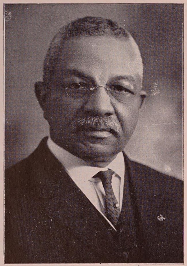 African American man wears a white shirt, dark tie, black jacket, and glasses