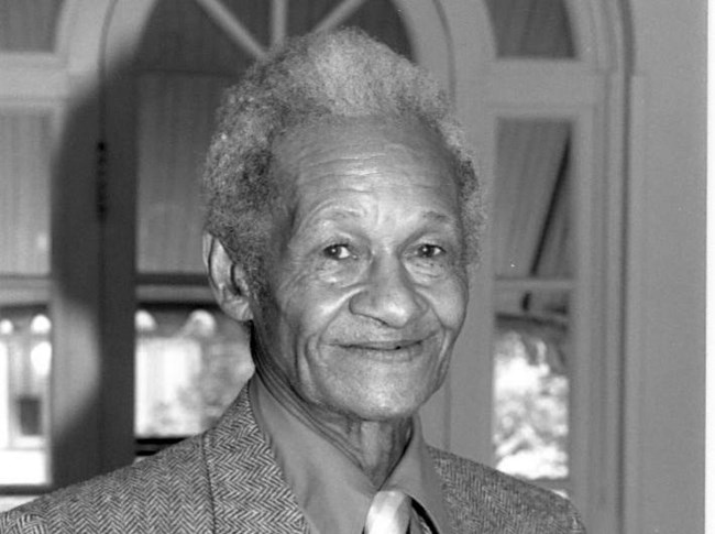 African American man with white hair stands indoors wearing a black and gray jacket, dark shirt, and striped tie.