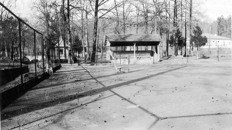 black and white photo of empty tennis courts surrounded by an iron fence with a bandstand facing it and trees and buildings in the background.