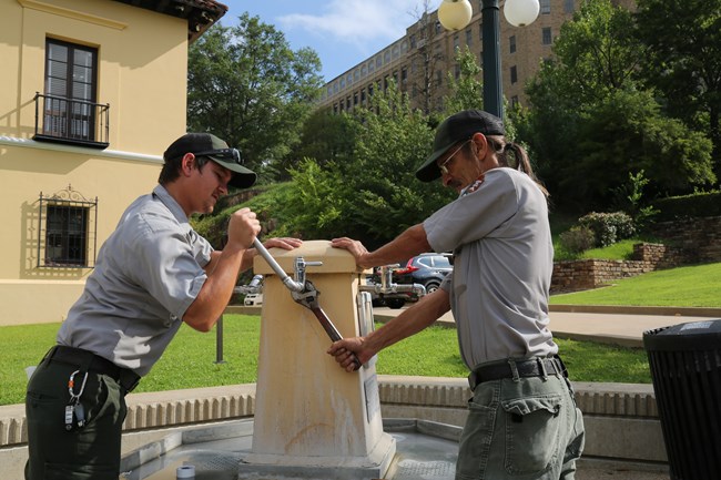Two men in gray shirts and green pants hold wrenches on a water fountain spigot.