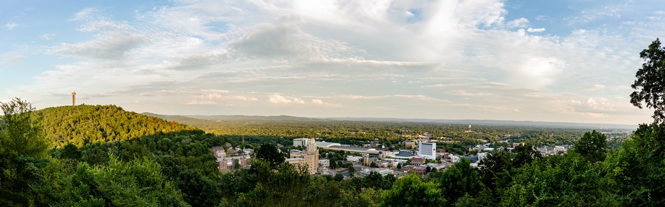 A panoramic view of downtown Hot Springs. A tower emerges from a rounded mountain on the left and buildings fill the valley below.