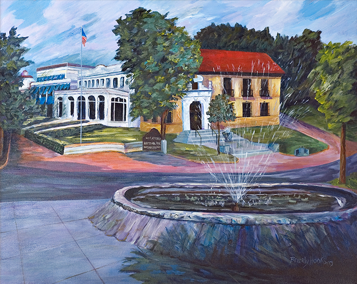 South end of Bathhouse Row from across the street. Foreground has a large round fountain with water spouting up from the center. Across the street is the park Administration building on the right, a Spanish style structure with yellowish stucco and a red tile roof and adjacent around the corner on the left is the Lamar Bathhouse, a white stucco structure.