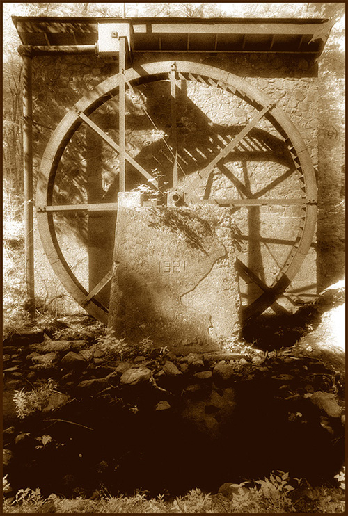 sepia tone photo of a stone structure with a large water wheel mounted on the side, shadowy foreground