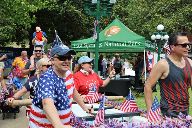 Four people push a decorated float during a parade as participants in the background are wearing red, white, and blue while walking past a park ranger tent.