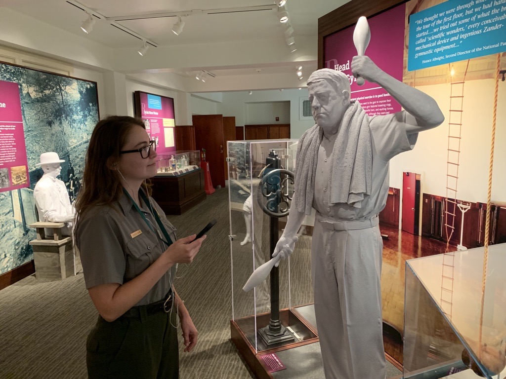 A woman wearing headphones holds a cellphone up to an exhibit with a white statue of a man.