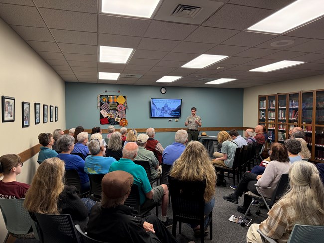 A crowd of people watch a ranger in a grey uniform speak in front of a presentation