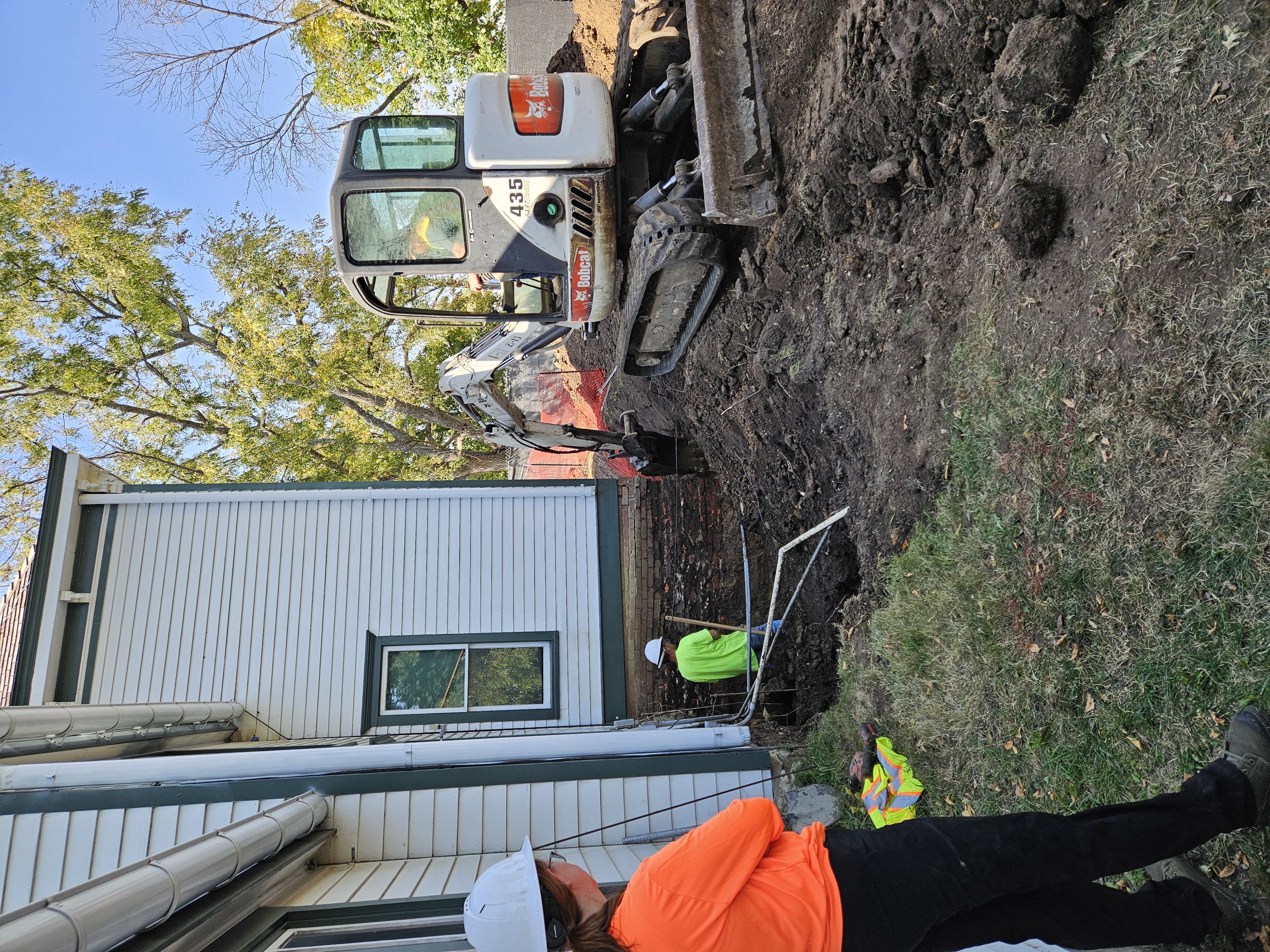 A woman (left) in the foreground looks on as a bob cat excavator (right) digs a trench around the farm home. A man (center) standing in the ditch monitors the digging.