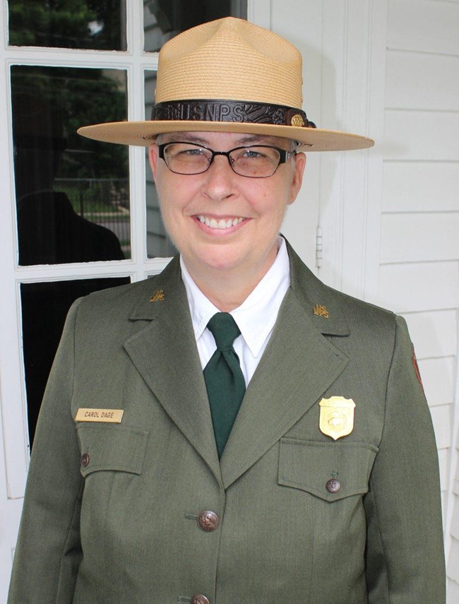Superintendent Carol J. Dage poses in front of the front door of the Truman Home, wearing the green Park Service uniform and straw Ranger hat. She is wearing glasses and smiling.