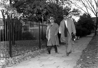 President Truman walking aside his home. He is wearing a hat and a long coat. A photographer is walking aside him. It appears as if it's an autumn season.