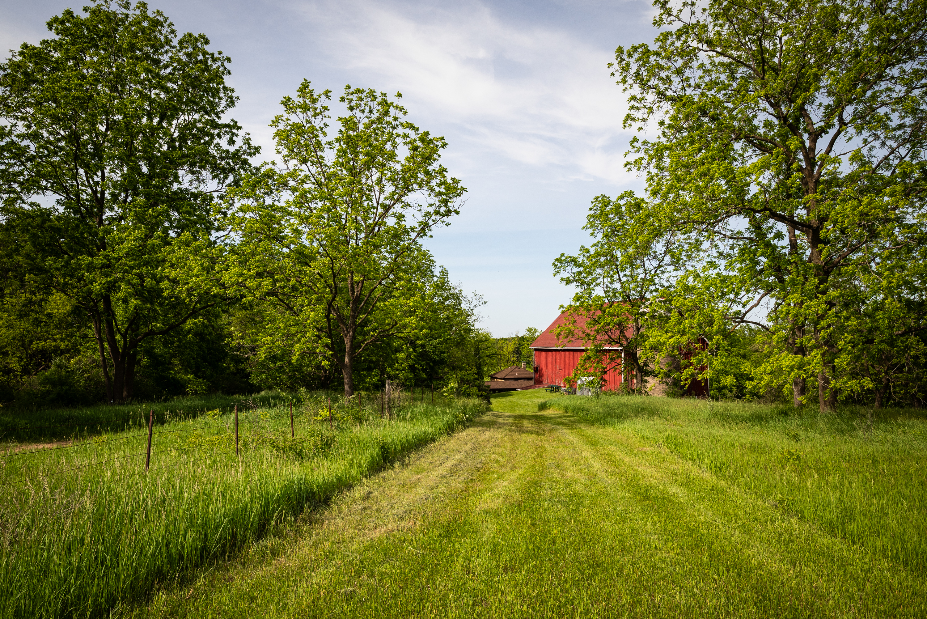 Photograph of a grassy trail with a red barn in the distance under a blue sky.