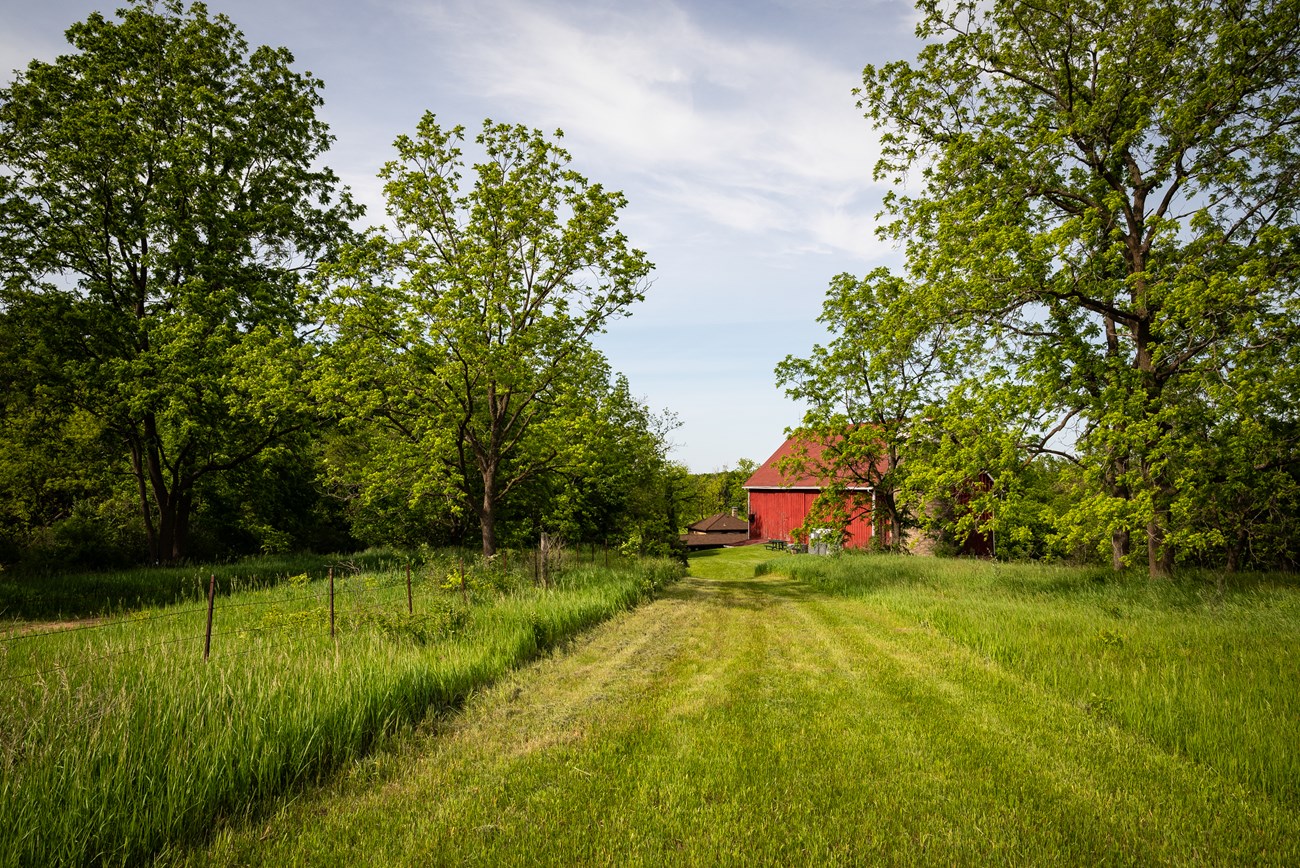 Photograph of a grassy trail with a red barn in the distance under a blue sky.