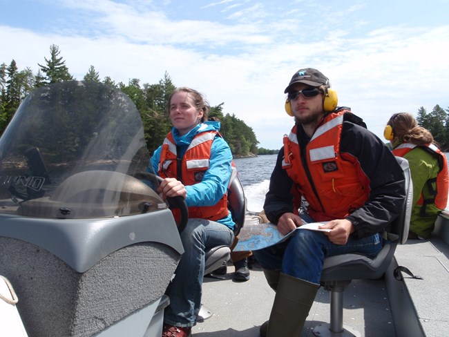 Three people sit in an open powerboat. The person in the back of the boat is looking away while the two in the front are looking ahead. A woman in a blue coat is driving the boat.